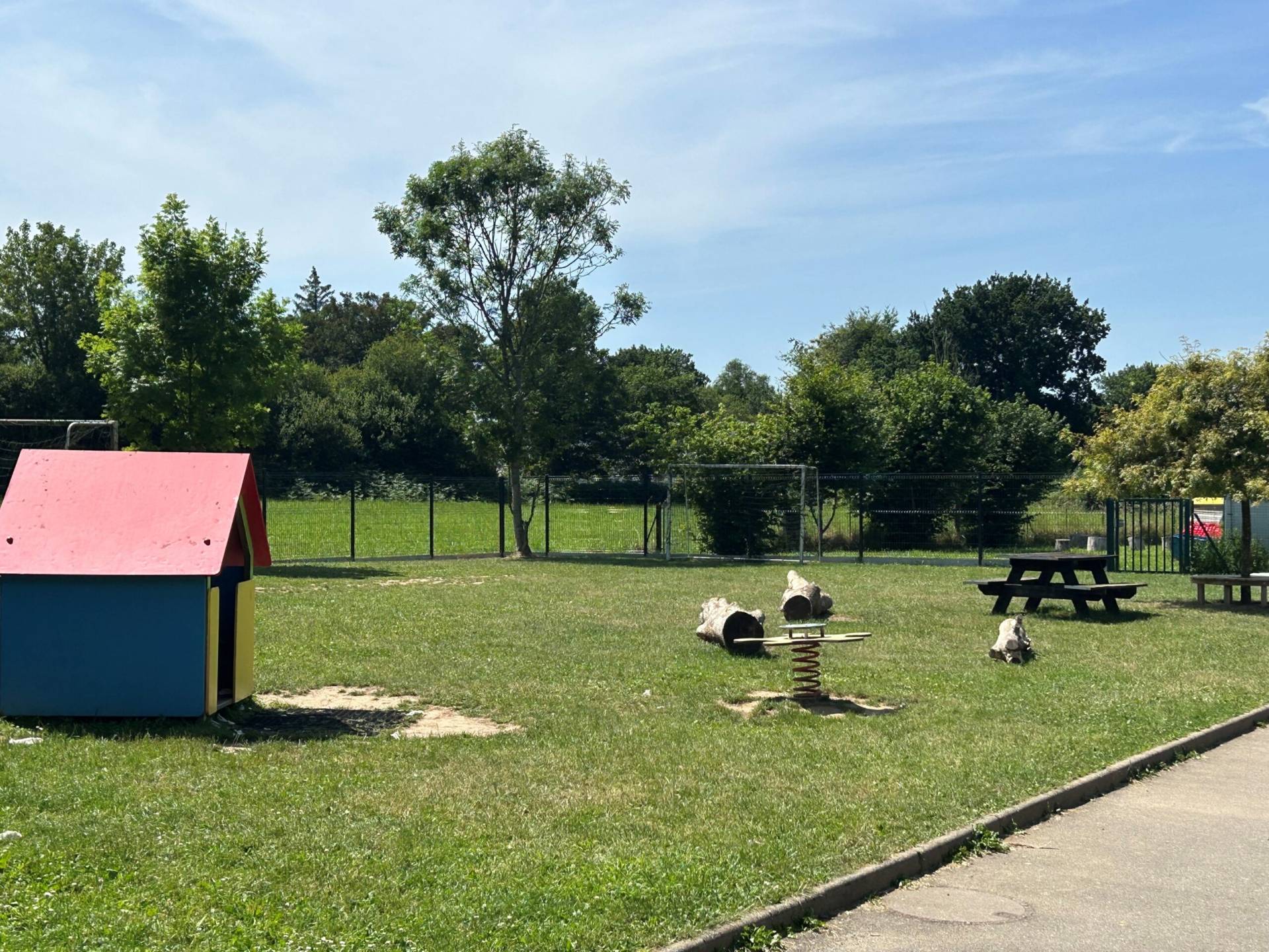 Colorful children's playhouse on a grassy field with trees and a fence in the background under a bright blue sky