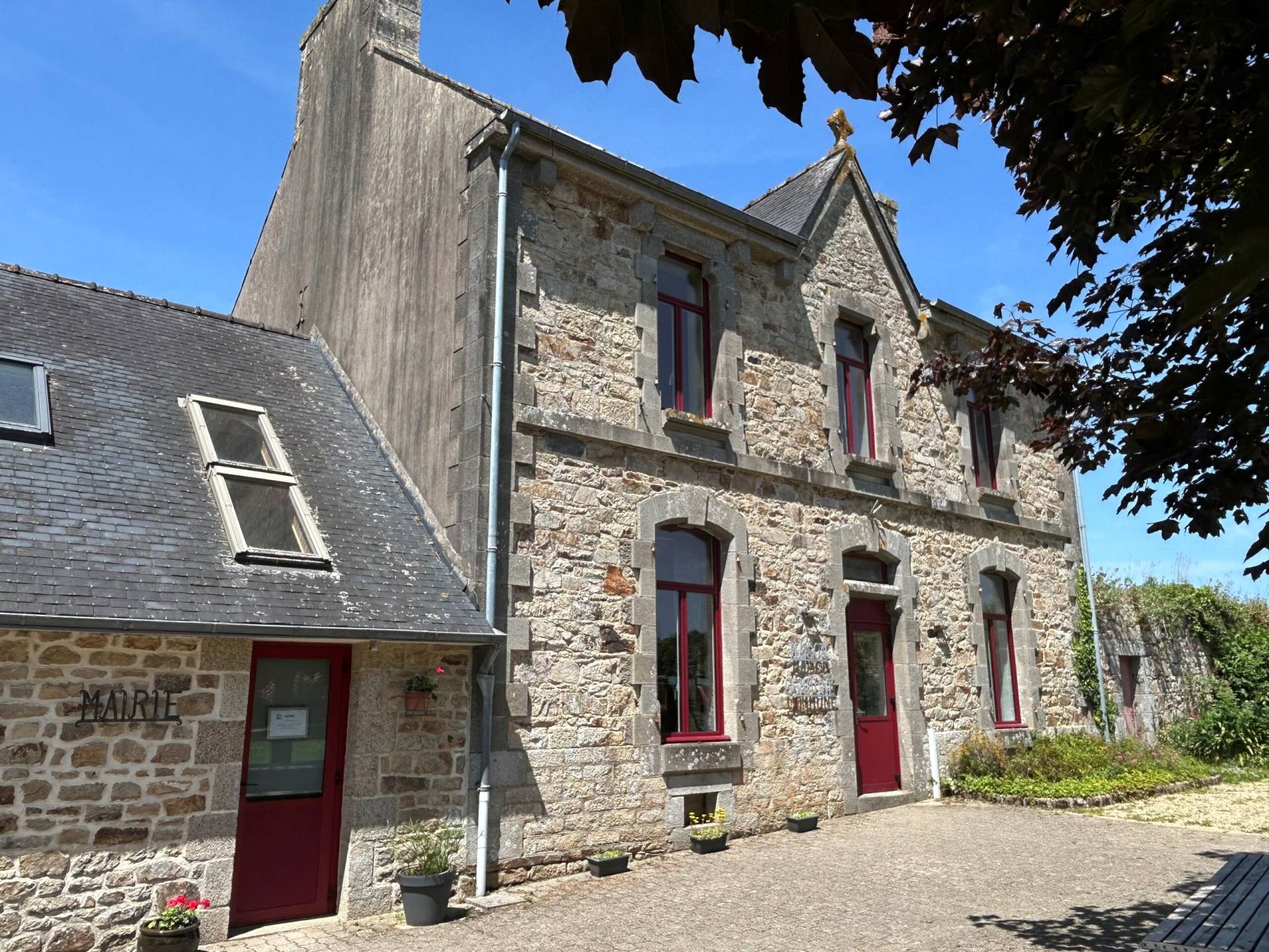 Stone town hall building with red-framed windows, slate roof, and potted plants along a sunny cobblestone courtyard.